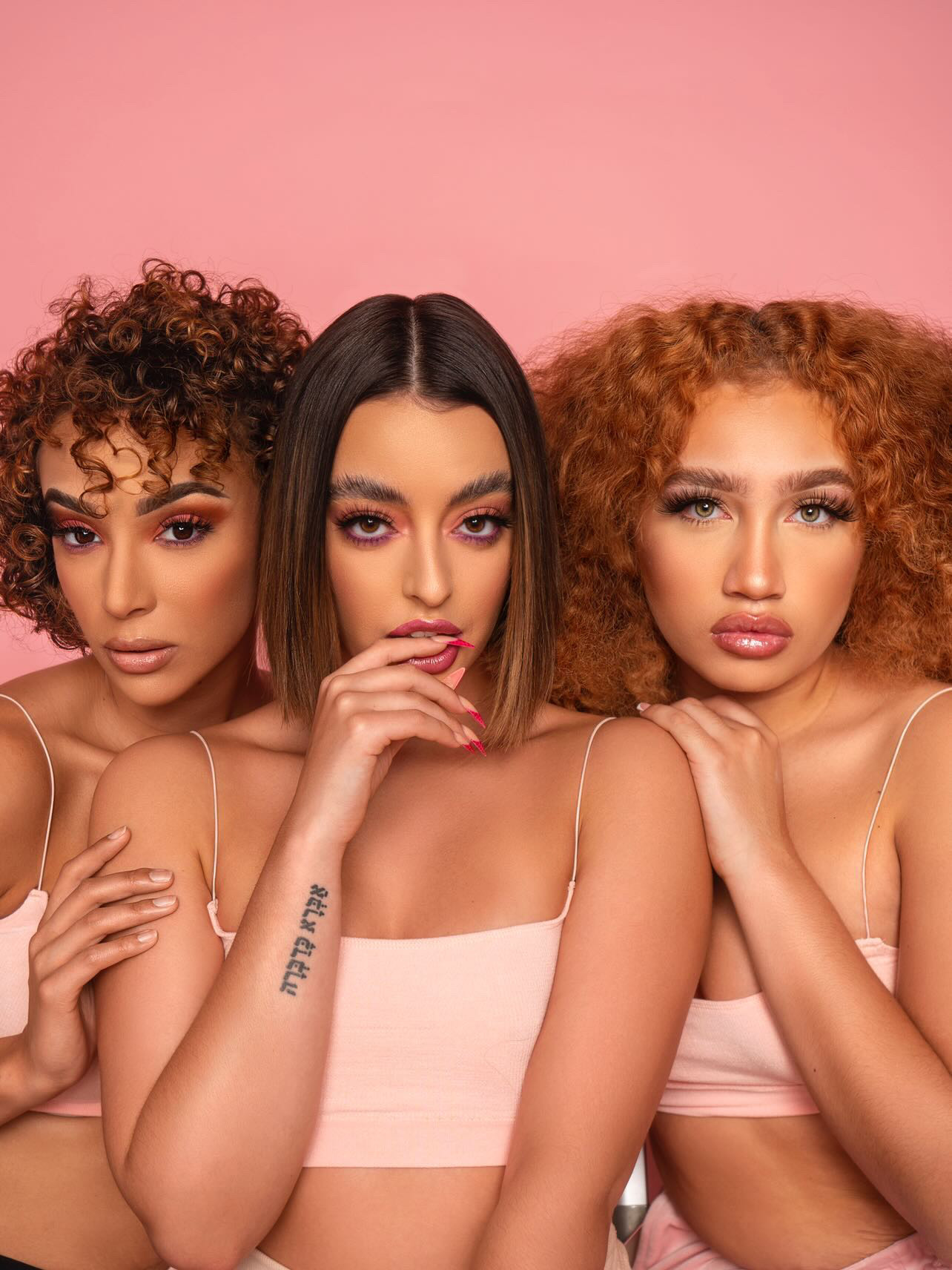 Three women with curly and straight hair pose against a pink background, wearing light-colored tops, with serious expressions.