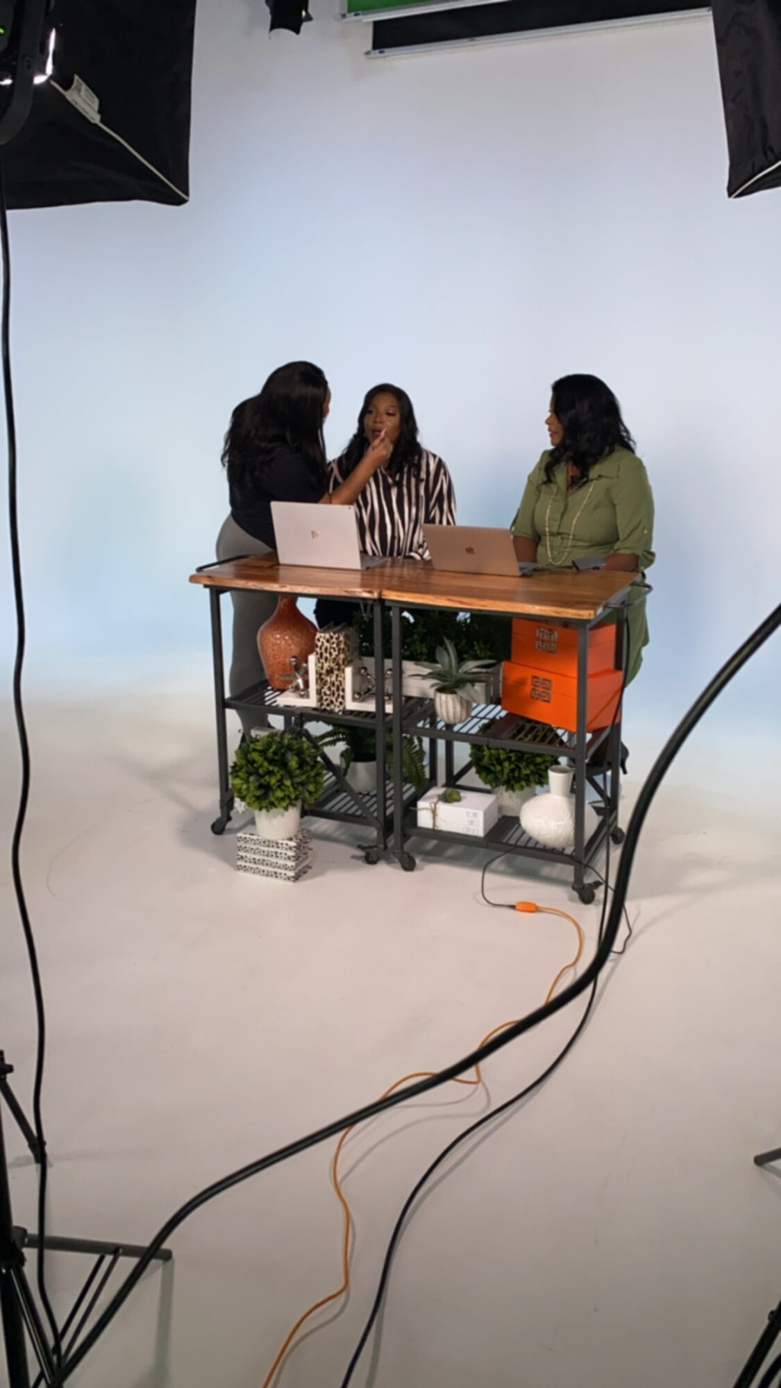 Three women sit at a table with a laptop in a studio with a white backdrop and equipment, viewed from a low angle.