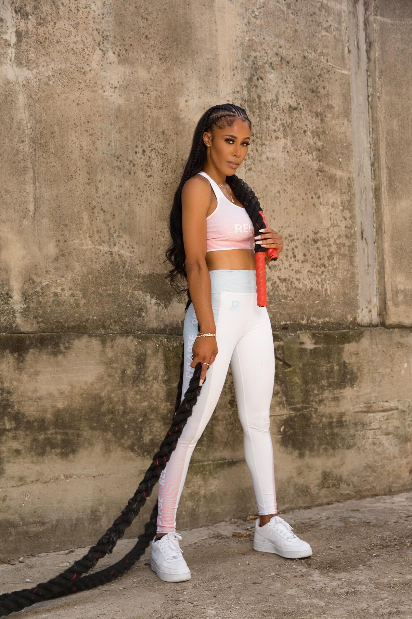Young woman with long braided hair in athletic outfit standing against a concrete wall, holding a water bottle.