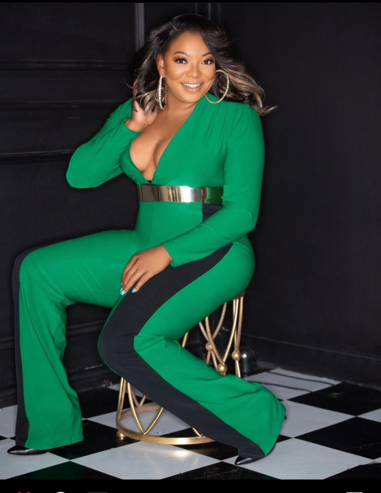 Woman in green jumpsuit sitting on a gold chair, smiling, black and white checkered floor, dark background.