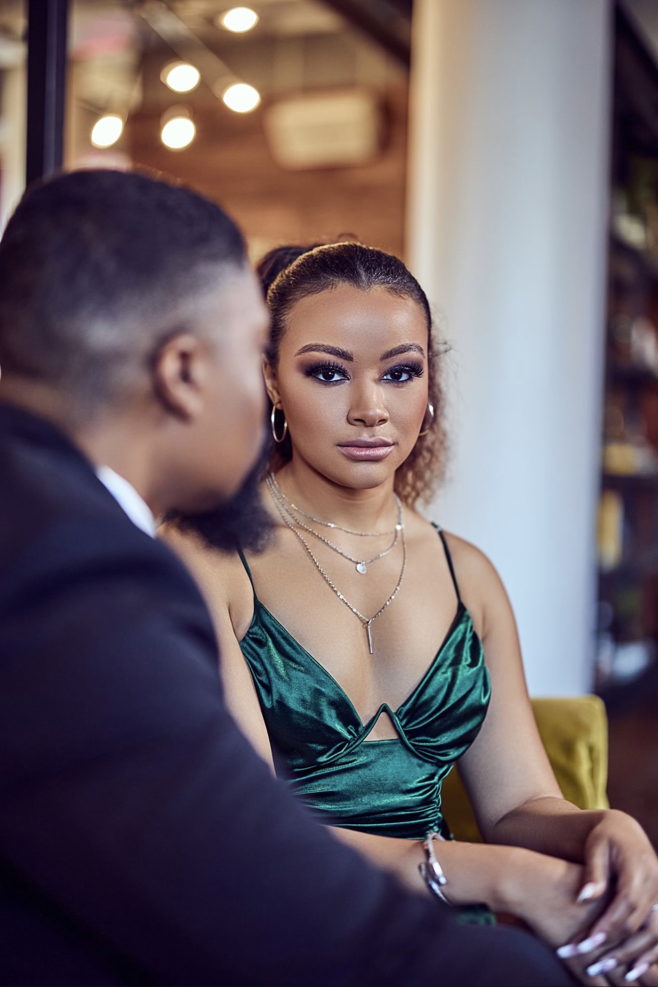 A woman with makeup and jewelry looks at a man in a suit, both seated indoors. The background is blurred.