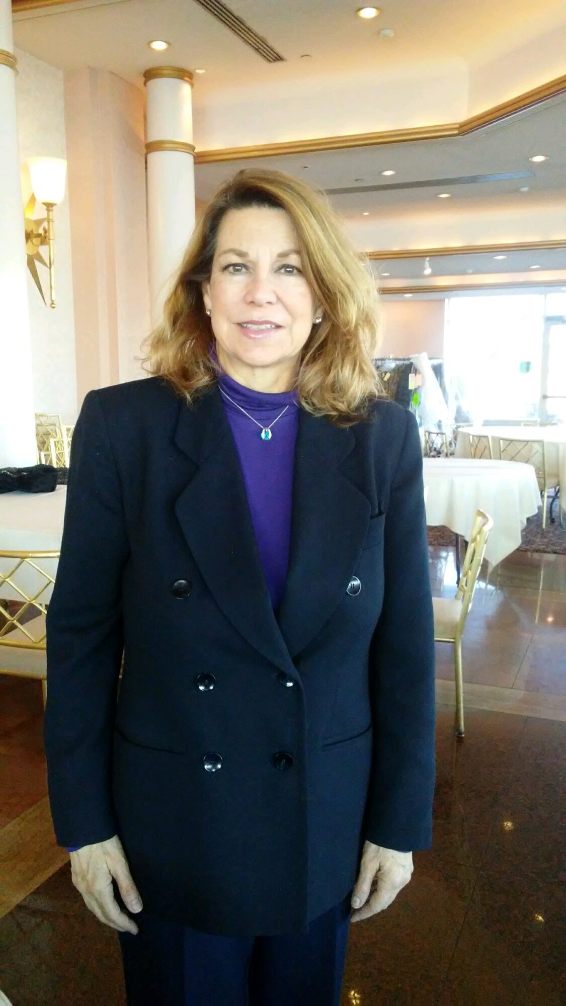 Woman with shoulder-length hair in a dark blazer and purple top standing indoors with elegant decor.