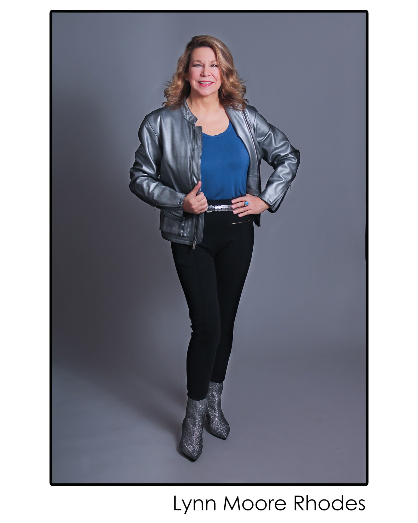 Woman with wavy hair wearing a blue top, silver jacket, black pants, and gray boots, standing with hands on hips.