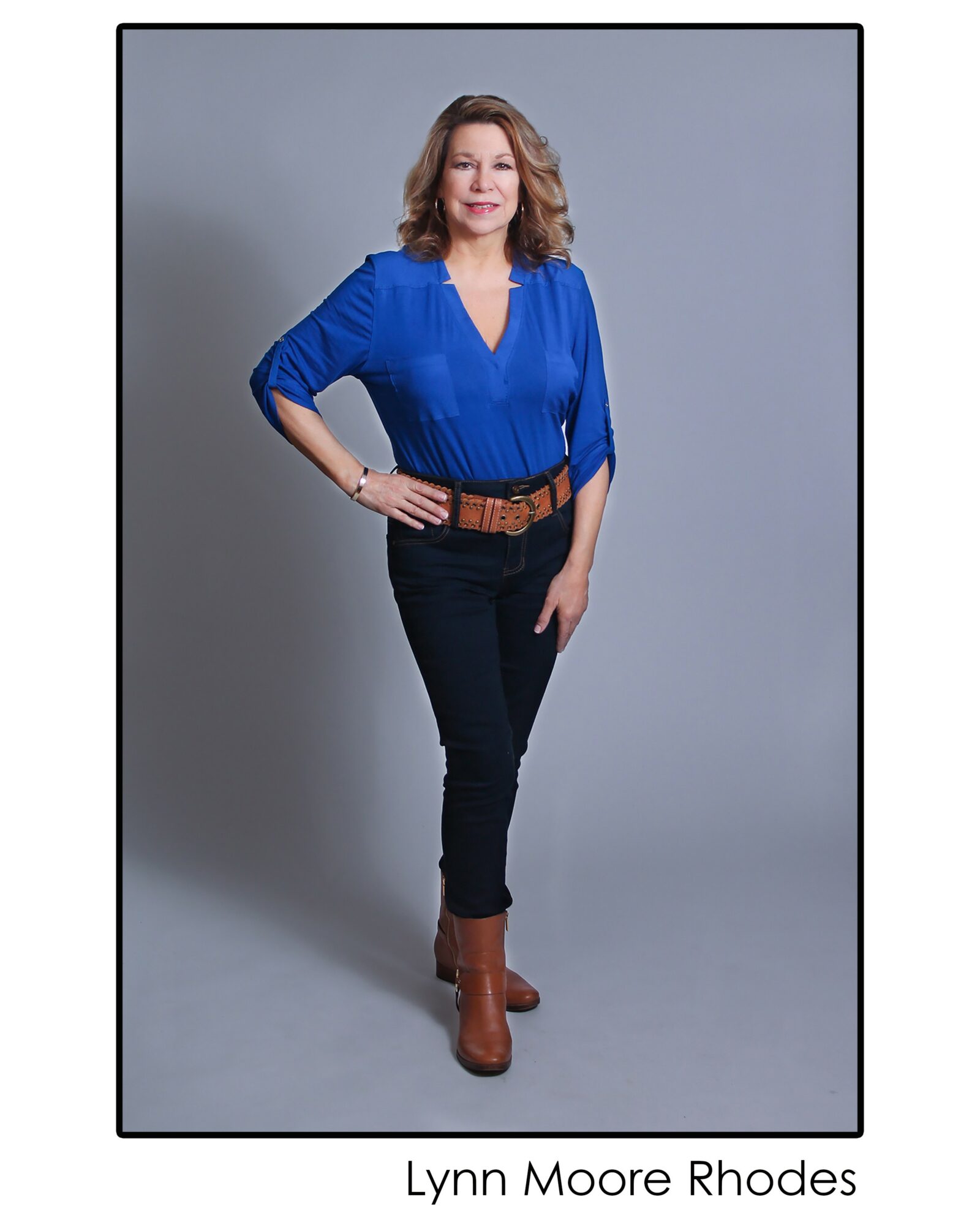 Woman with shoulder-length hair wearing a blue blouse, black pants, and brown boots, standing with one hand on her hip.