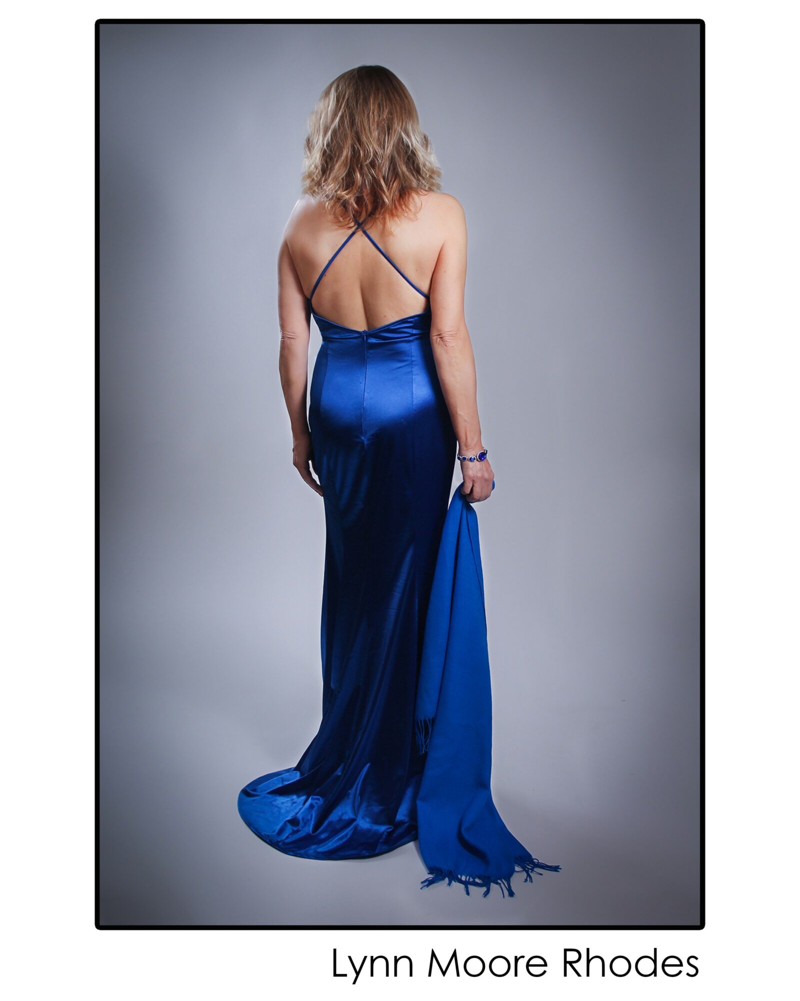 Woman in a long blue dress with spaghetti straps, holding the dress's train, facing away from the camera.