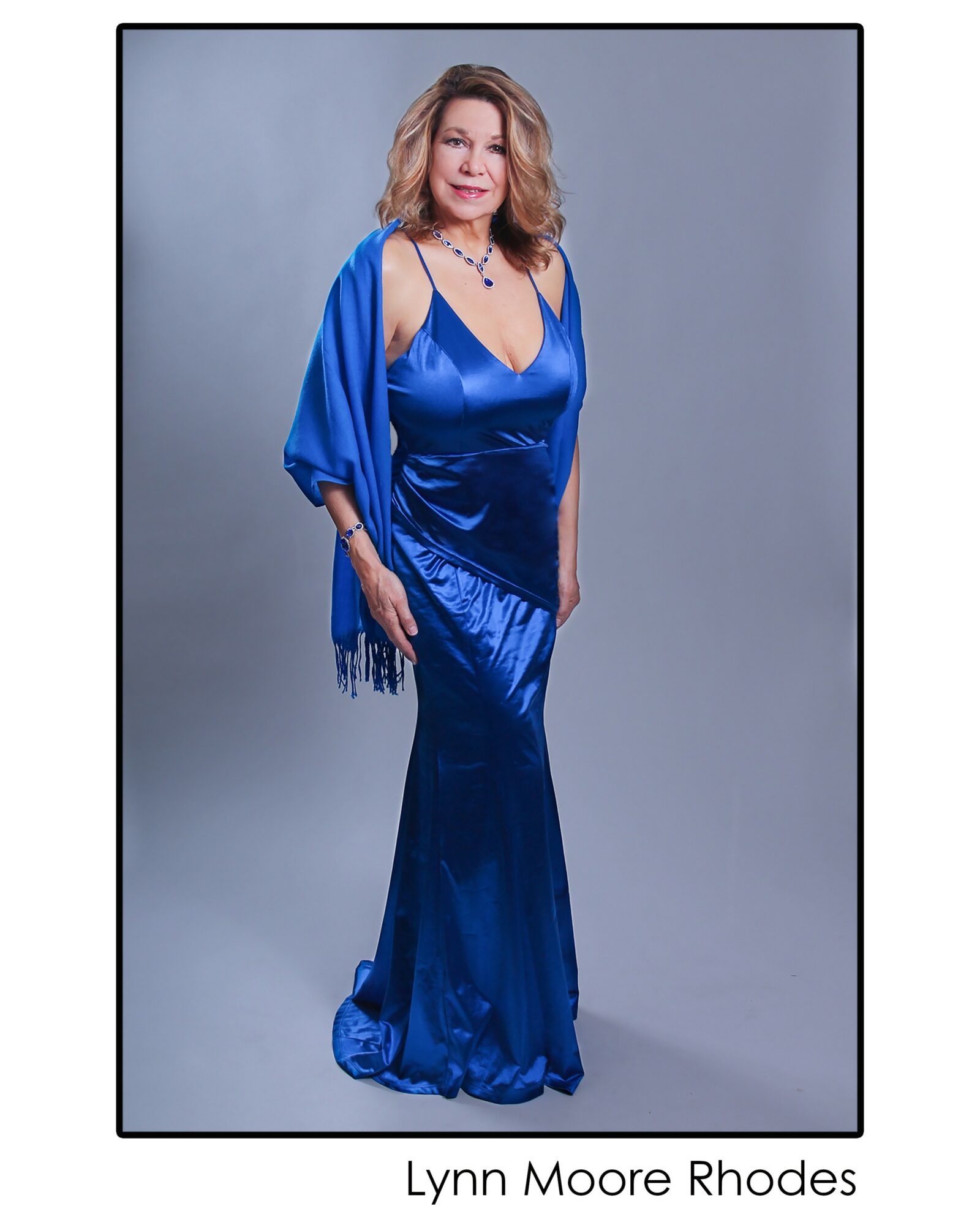Woman in a long, shiny blue dress with a matching shawl, standing against a gray background.