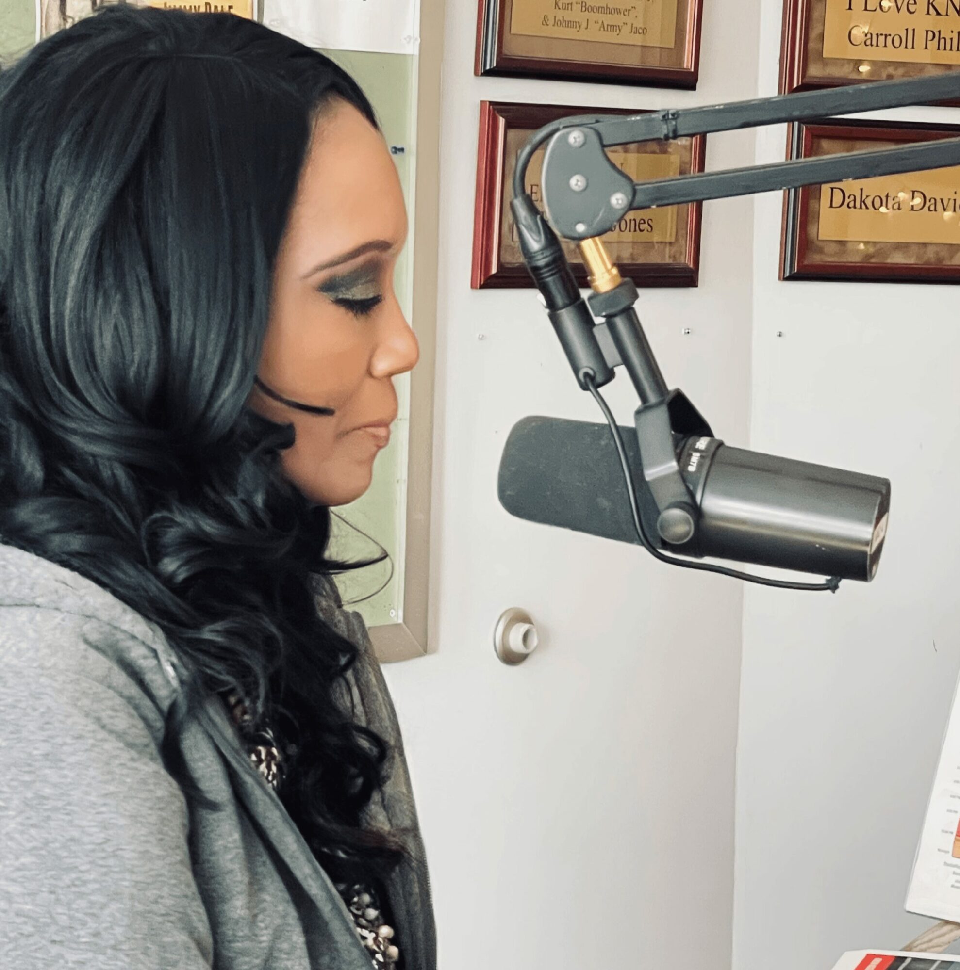 Woman with dark hair speaking into a professional microphone in a recording studio, framed certificates on the wall.
