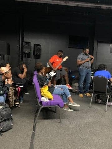 Group of people in a room, some sitting and some standing, with a person playing guitar and another speaking into a microphone.