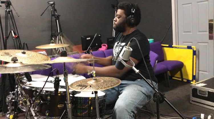 Person playing drums in a music studio with recording equipment and colorful furniture, wearing headphones.