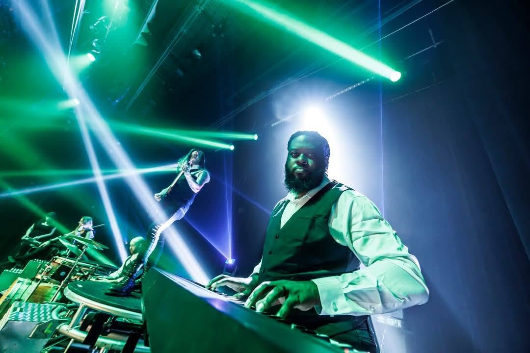 A DJ with a beard and glasses stands behind a turntable setup on stage, illuminated by colorful laser lights and a bright backlight.