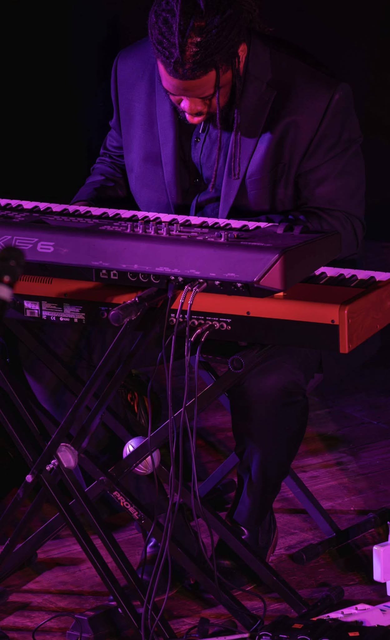 Person playing keyboard on stage with purple lighting, wearing headphones, focused on music, with cables and equipment visible.