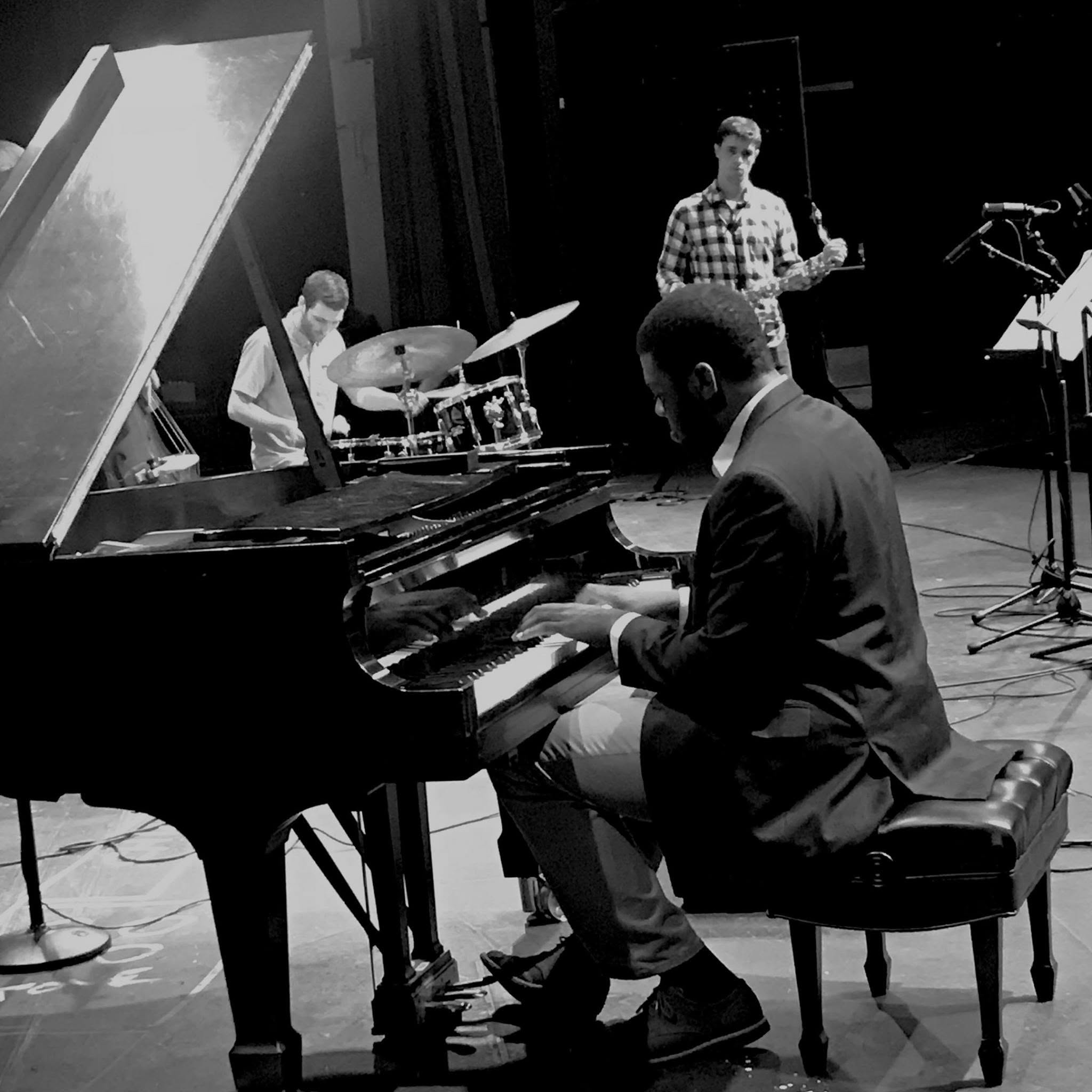 Three musicians perform on stage: a pianist, a drummer, and a guitarist, in black and white. Word count: 20.