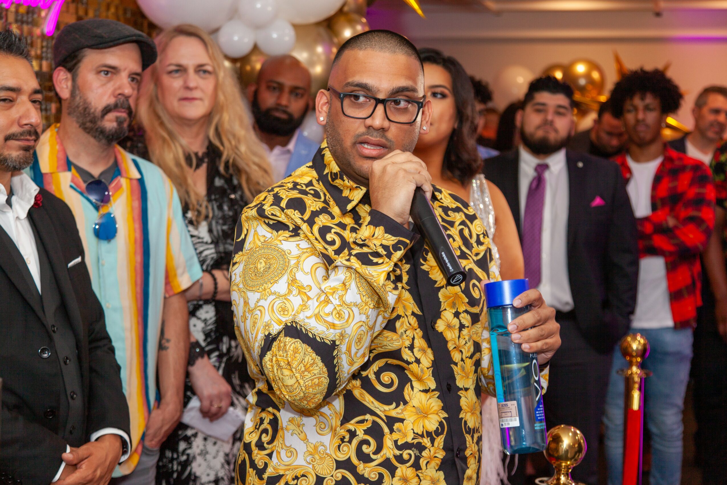 Man in glasses and ornate gold and black patterned shirt holding a microphone at a celebration with a group of people behind him.