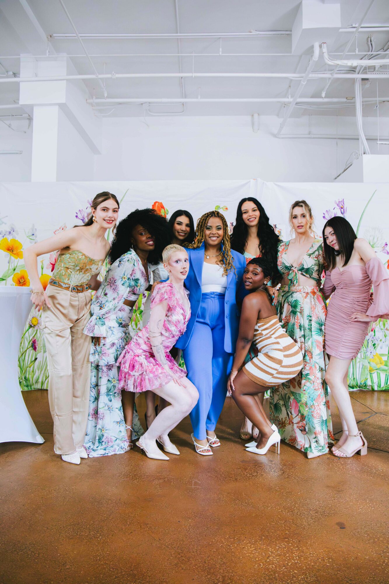 Group of women in colorful dresses posing indoors with a white wall and floral decorations behind them.