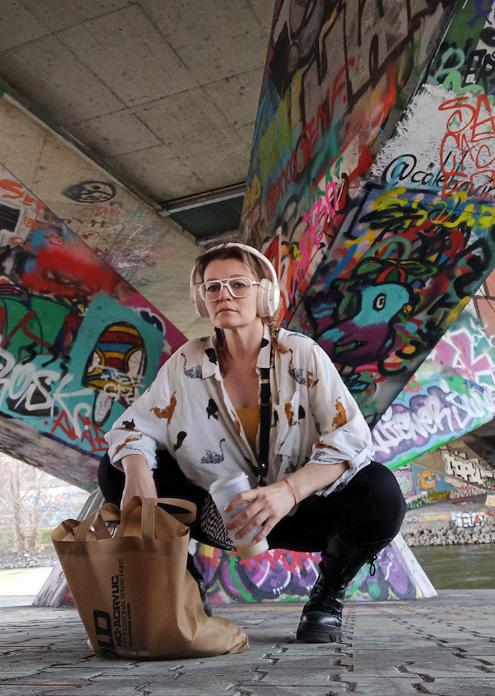 Person with glasses and headphones crouching under a graffiti-covered bridge, holding a paper bag, with colorful graffiti behind.
