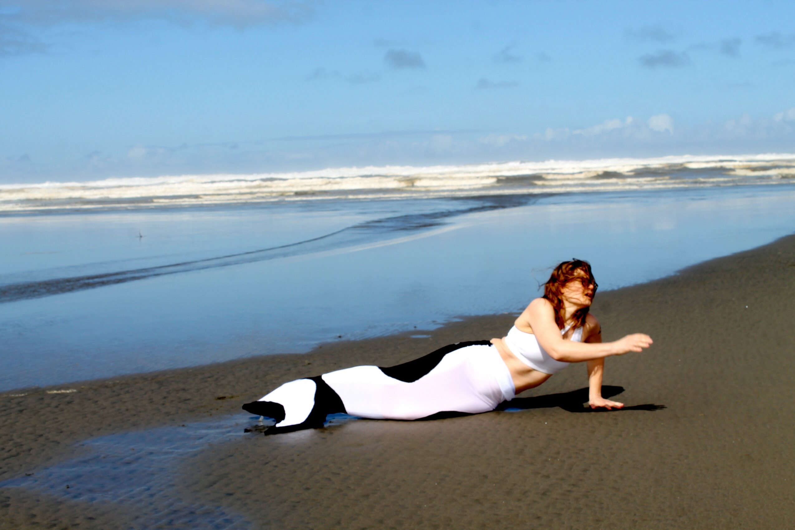 Person lying on beach sand, reaching forward with one arm, ocean waves in background, clear sky, and water reflecting the sky.