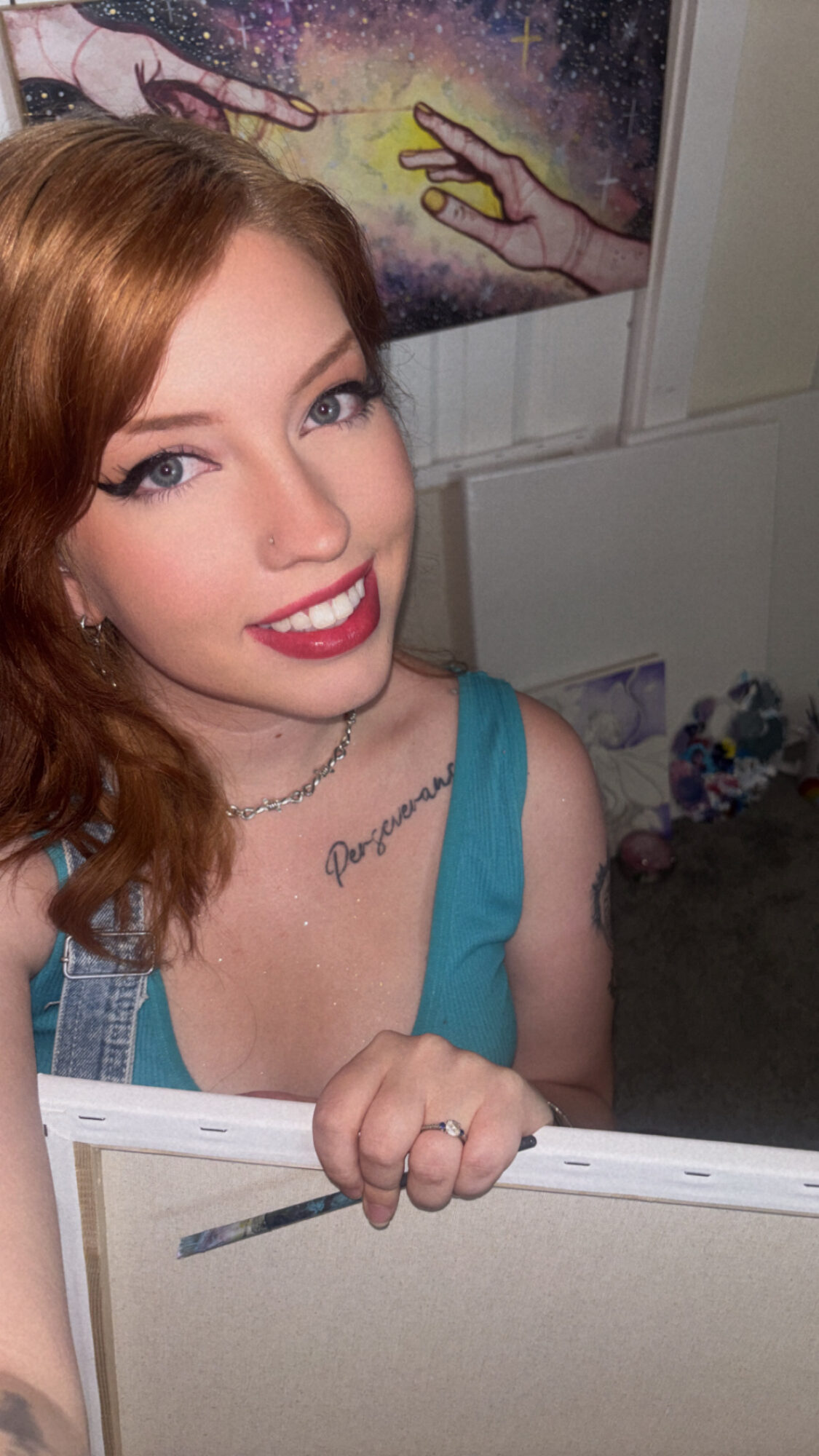 Young woman with red hair, blue top, smiling, holding a white object, background with colorful artwork.