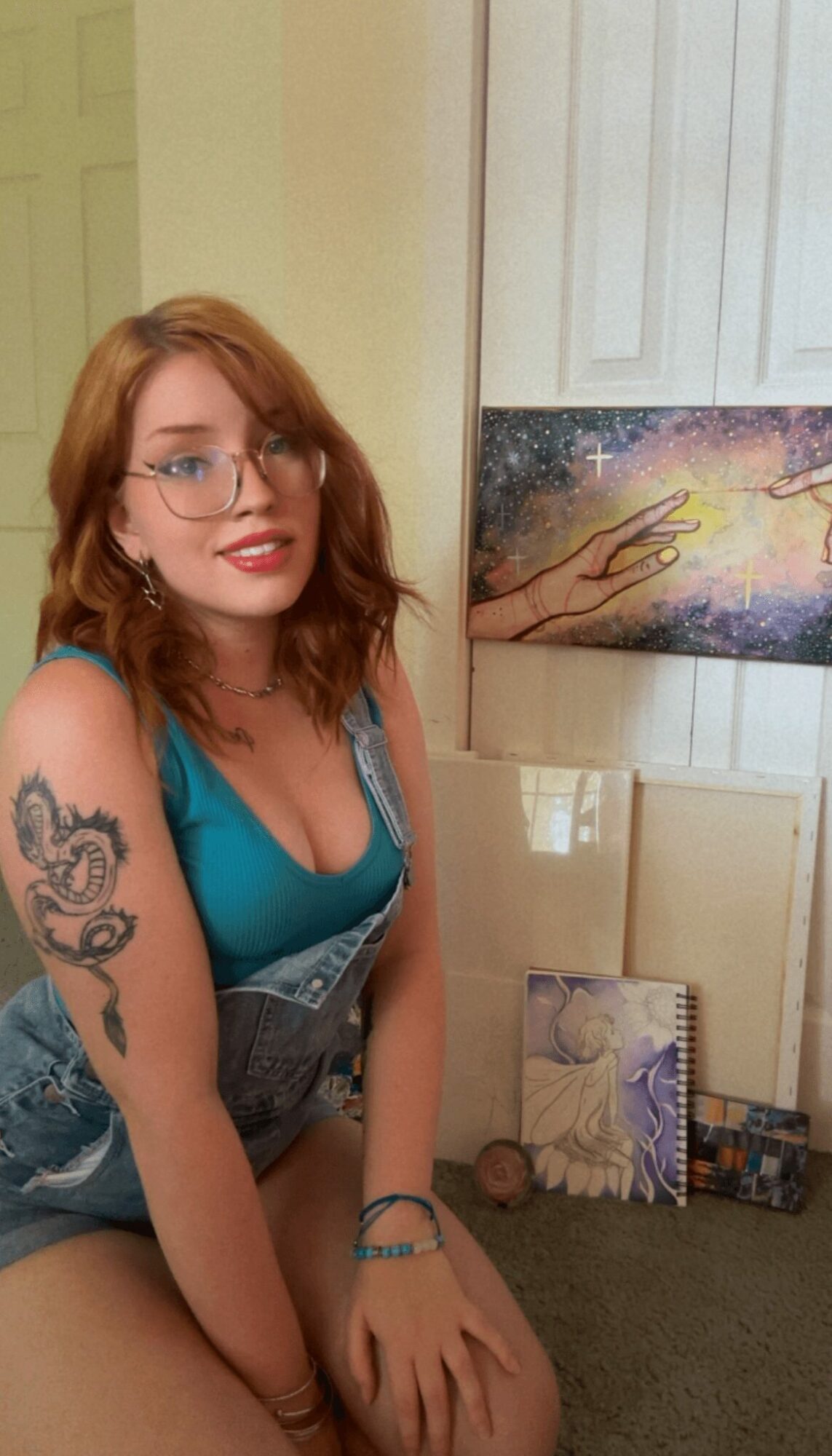 Young woman with glasses and red hair, wearing a teal top, sitting indoors with artwork behind her.