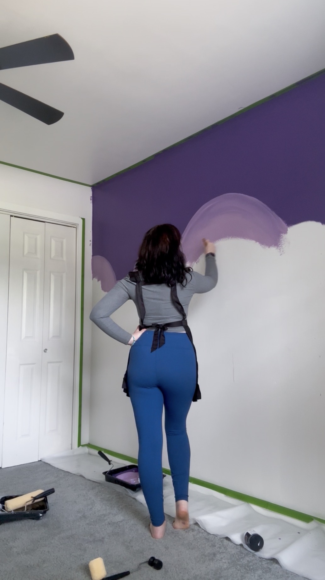 Person painting a purple wall white with a brush, standing on a floor covered with a drop cloth.