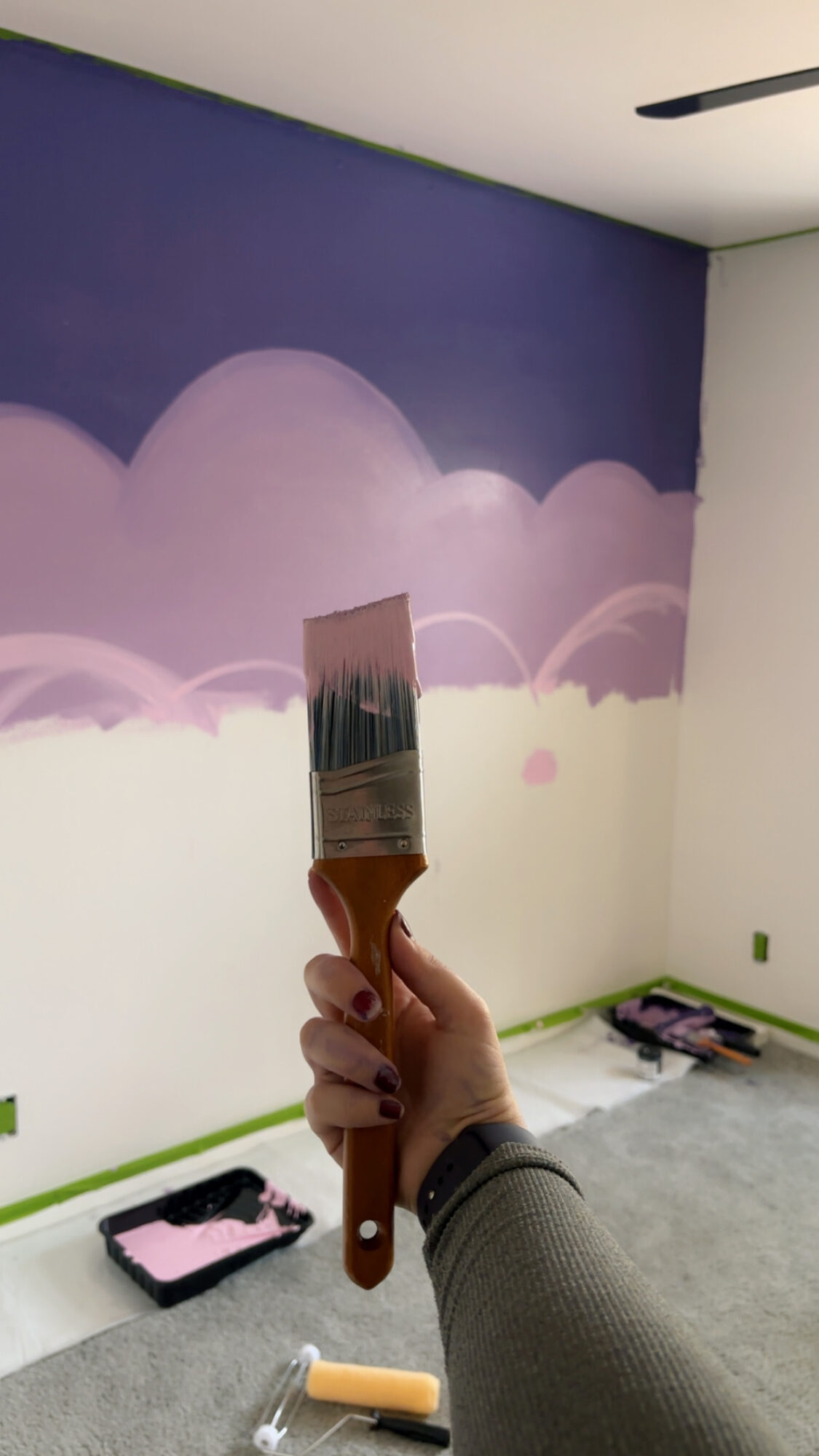 Person painting a wall with purple and pink clouds, holding a paintbrush, in a room with painting supplies on the floor.