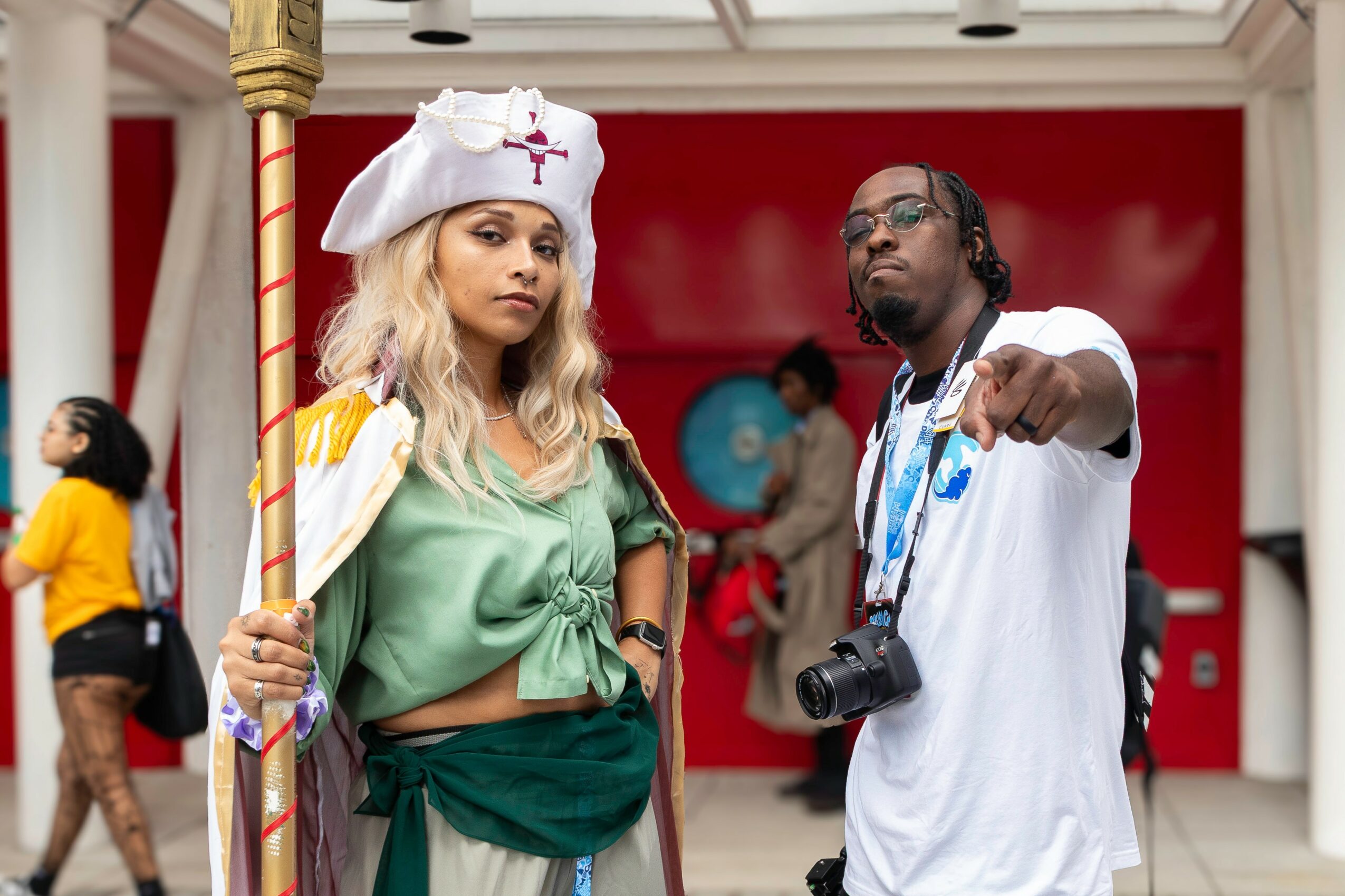 Two people pose indoors, one holding a staff, the other with a camera, with a red background and other people in the background.