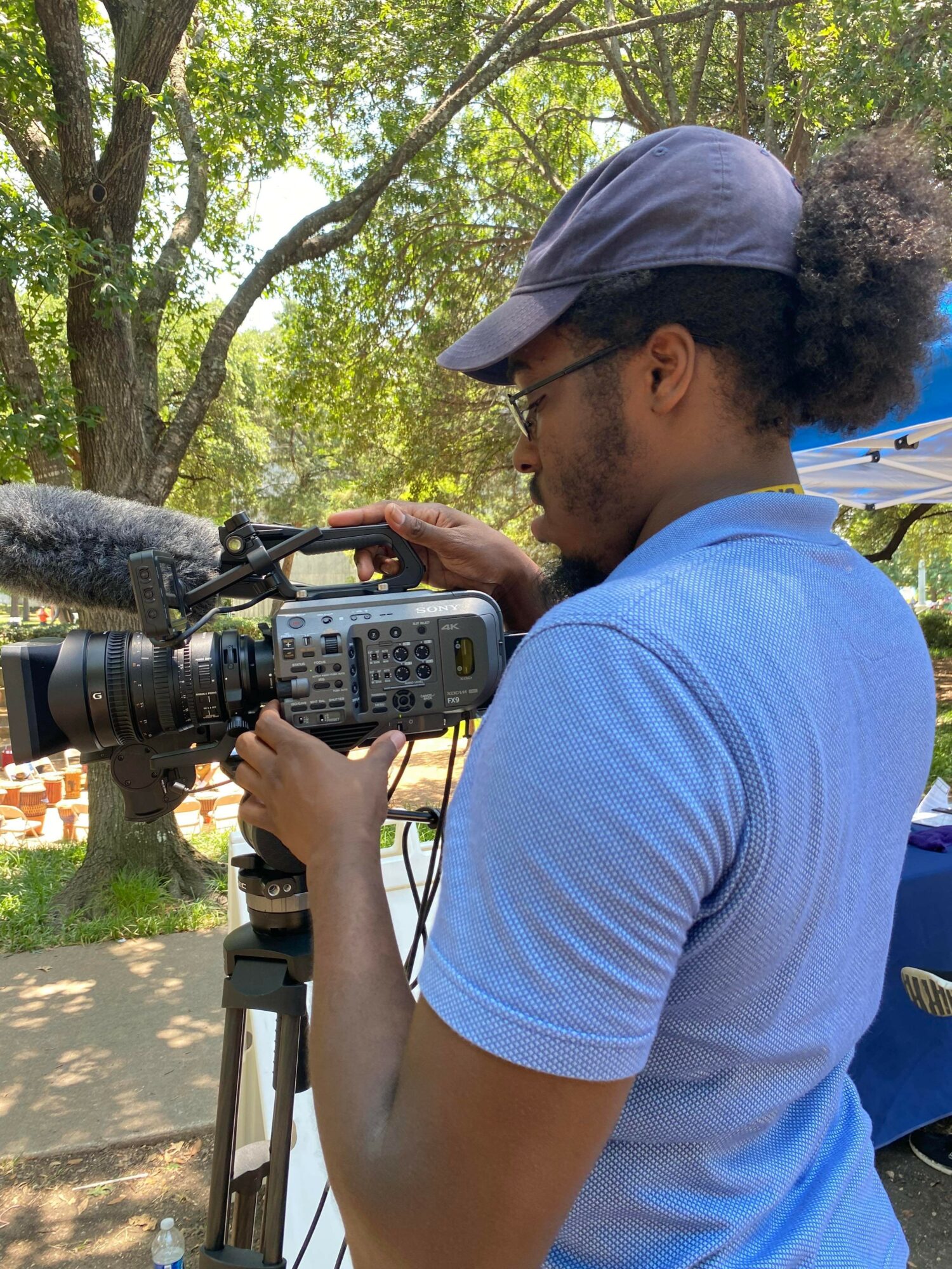 Person operating a professional video camera outdoors, wearing a blue shirt and cap, with trees in the background.