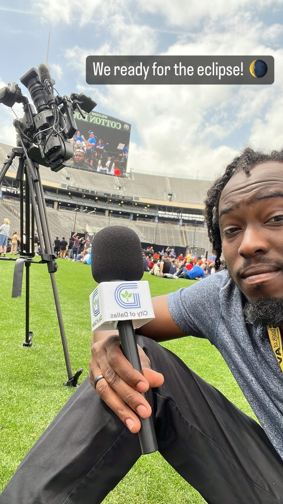 Person holding a microphone at a stadium during an eclipse, with a camera on a tripod and a scoreboard in the background.