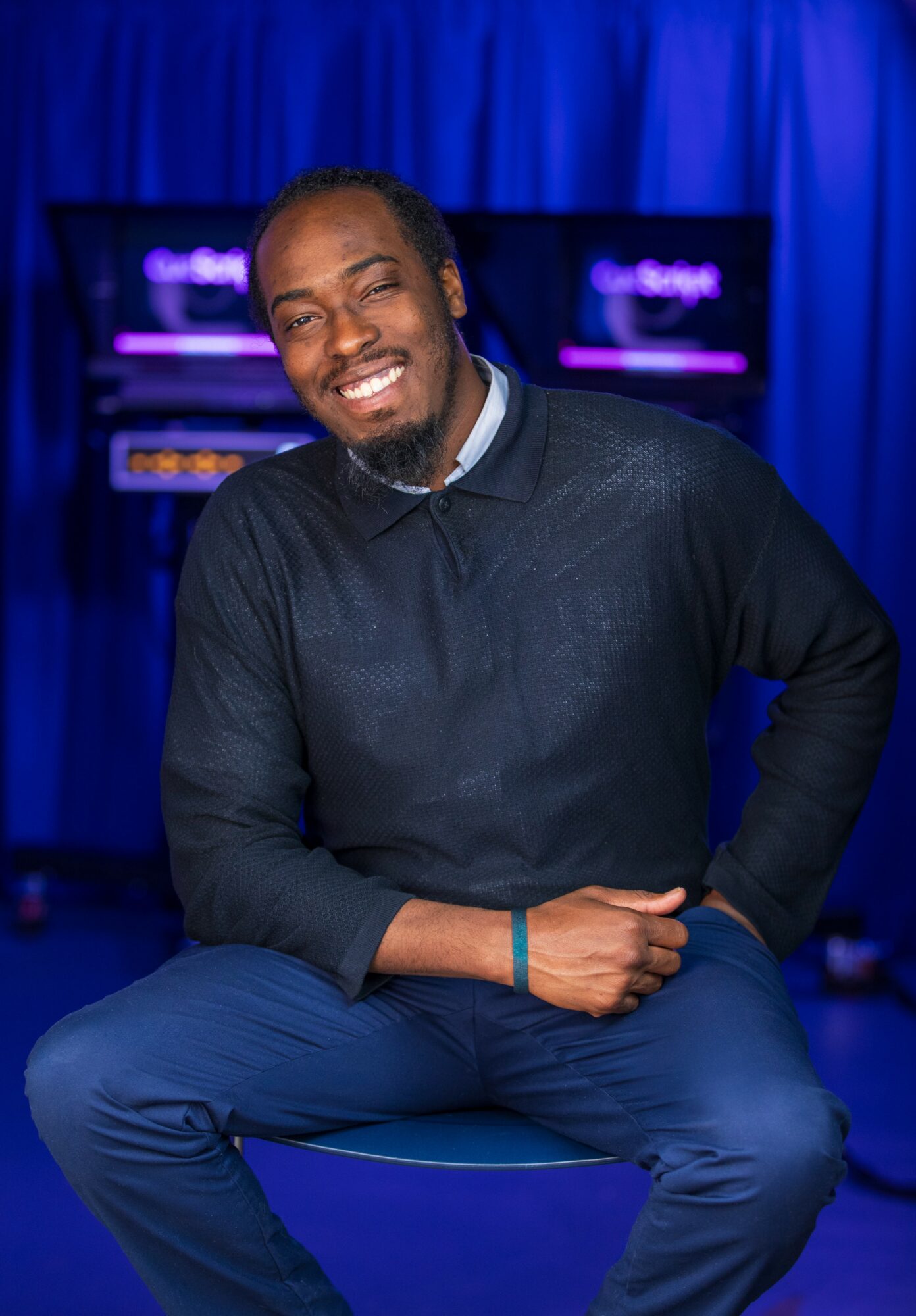Smiling man sitting on a stool with a blue curtain background and screens behind him.