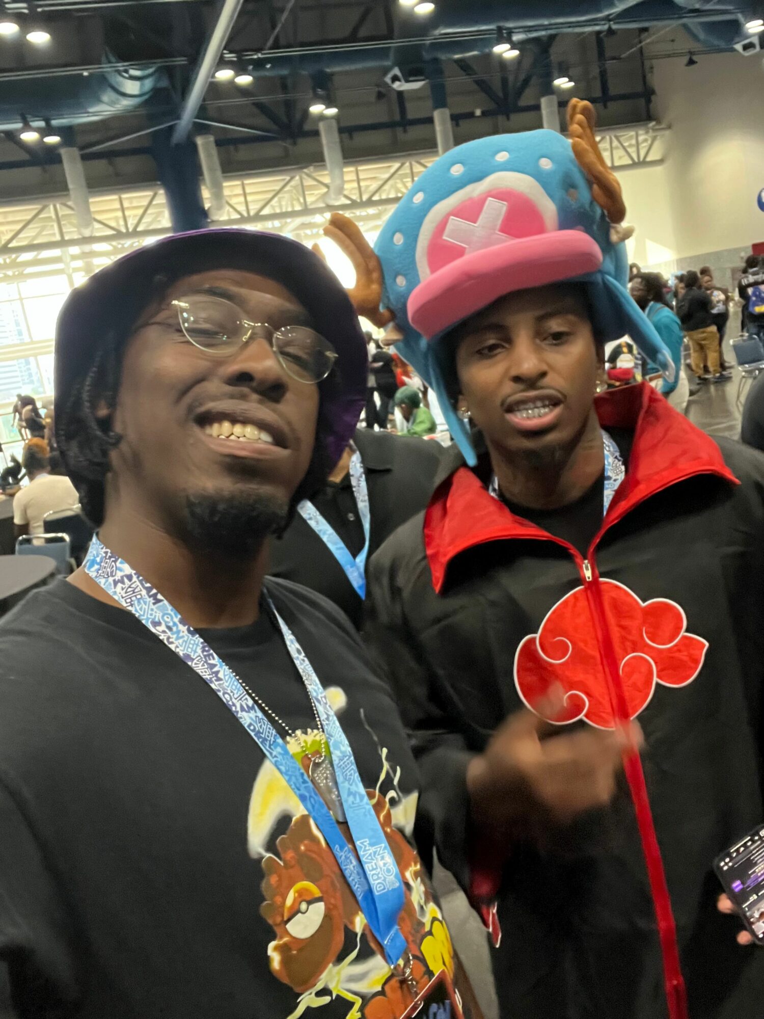 Two people in costumes smiling at a convention, one with a pink hat and the other with glasses and a lanyard.