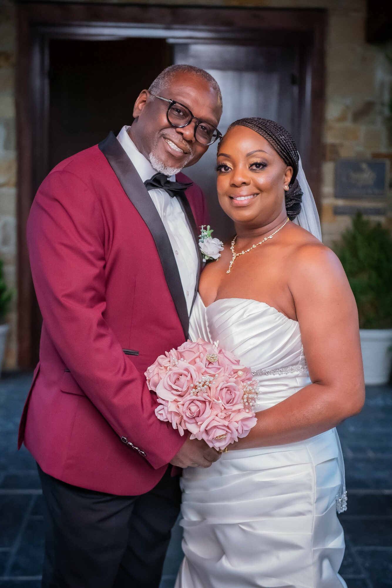 A bride and groom pose together indoors, the bride holding a pink bouquet, both smiling, dressed in wedding attire.