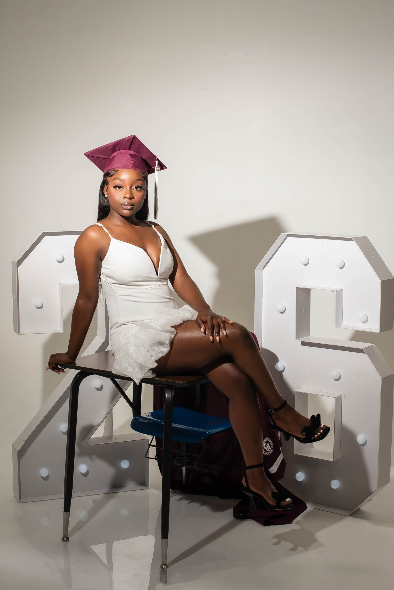 Young woman in a white dress and graduation cap, sitting on chairs, with large decorative letters behind her.