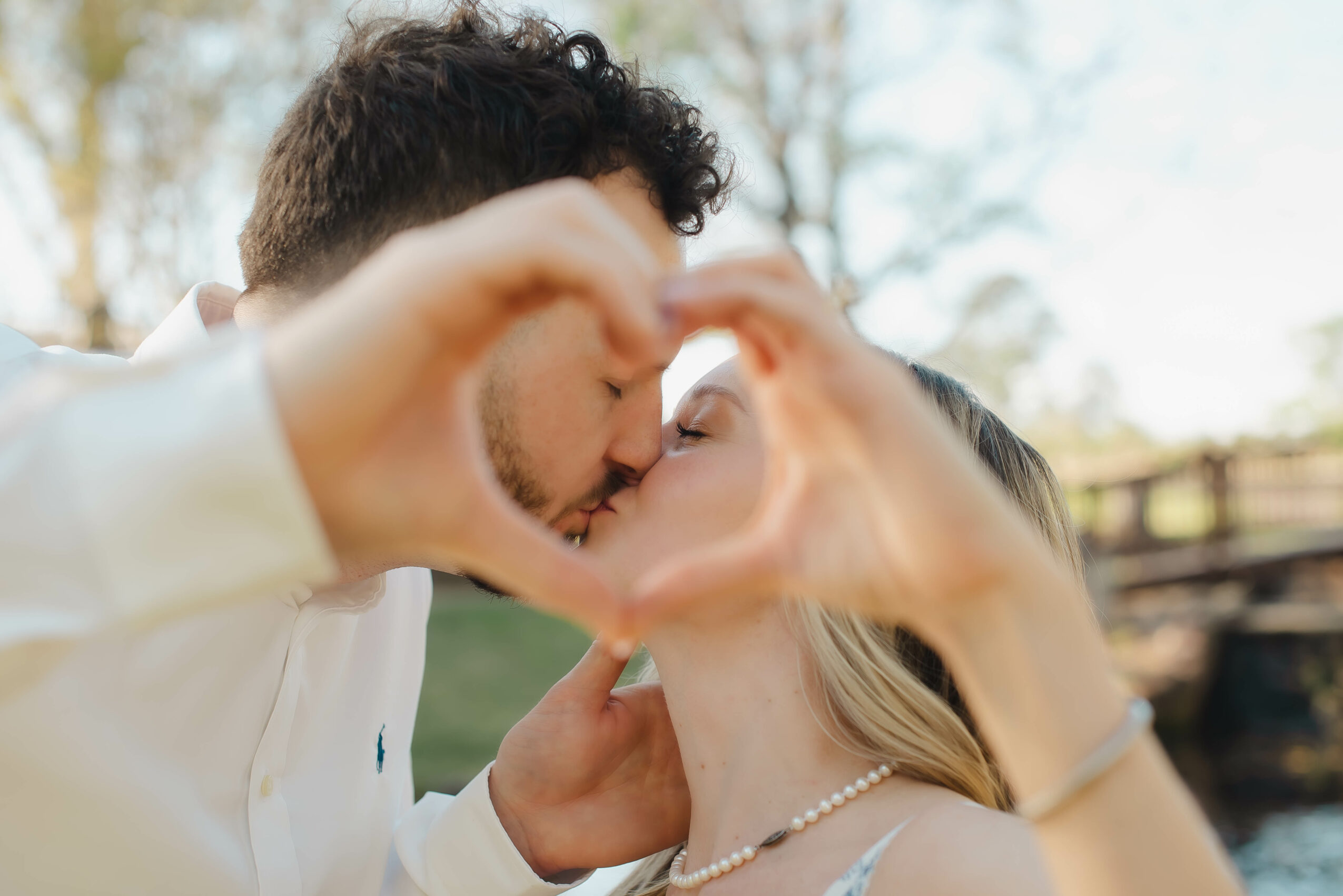 Two people kiss outdoors, forming a heart shape with their hands around their faces.