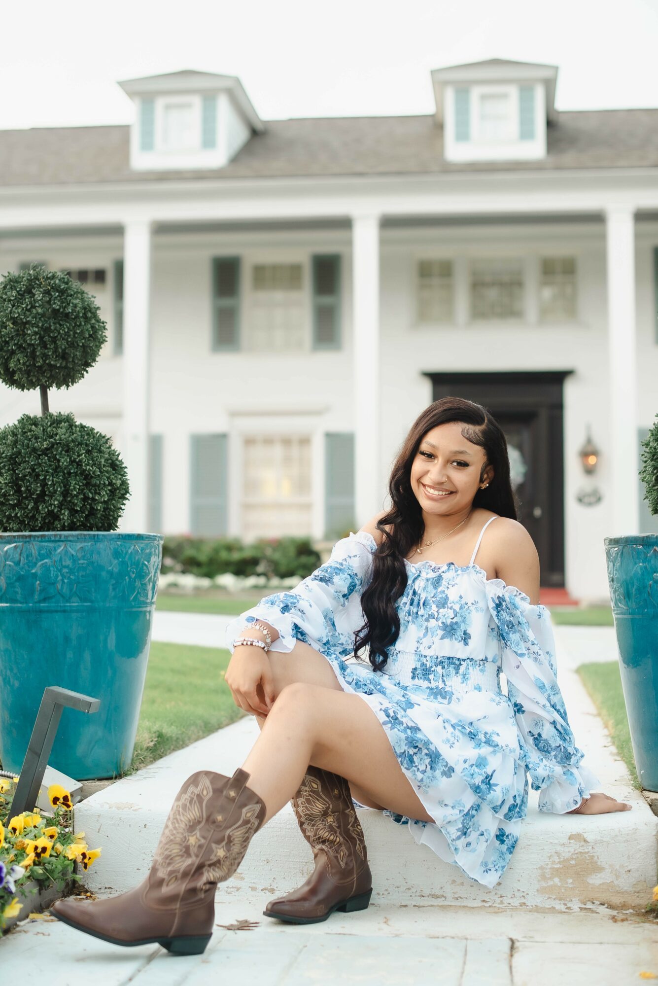 Young woman sitting outdoors in front of a large white house, smiling, wearing a blue floral dress and cowboy boots.
