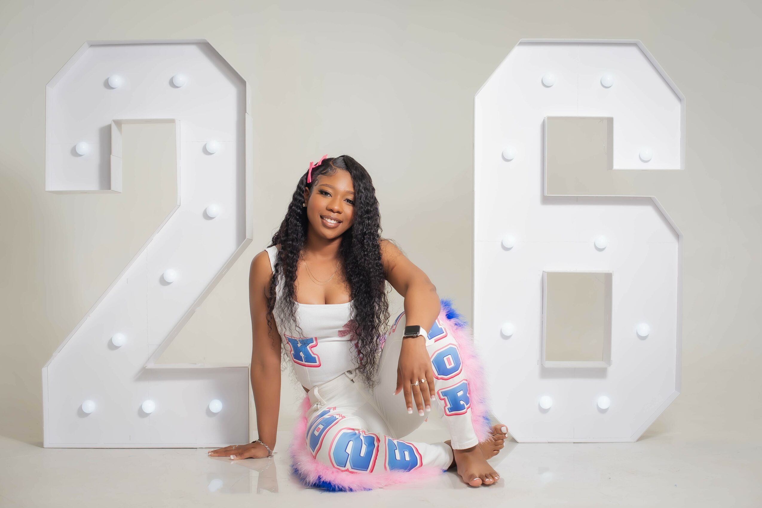 Young girl with long curly hair sitting between large white number signs 2 and 6, smiling, on a plain background.