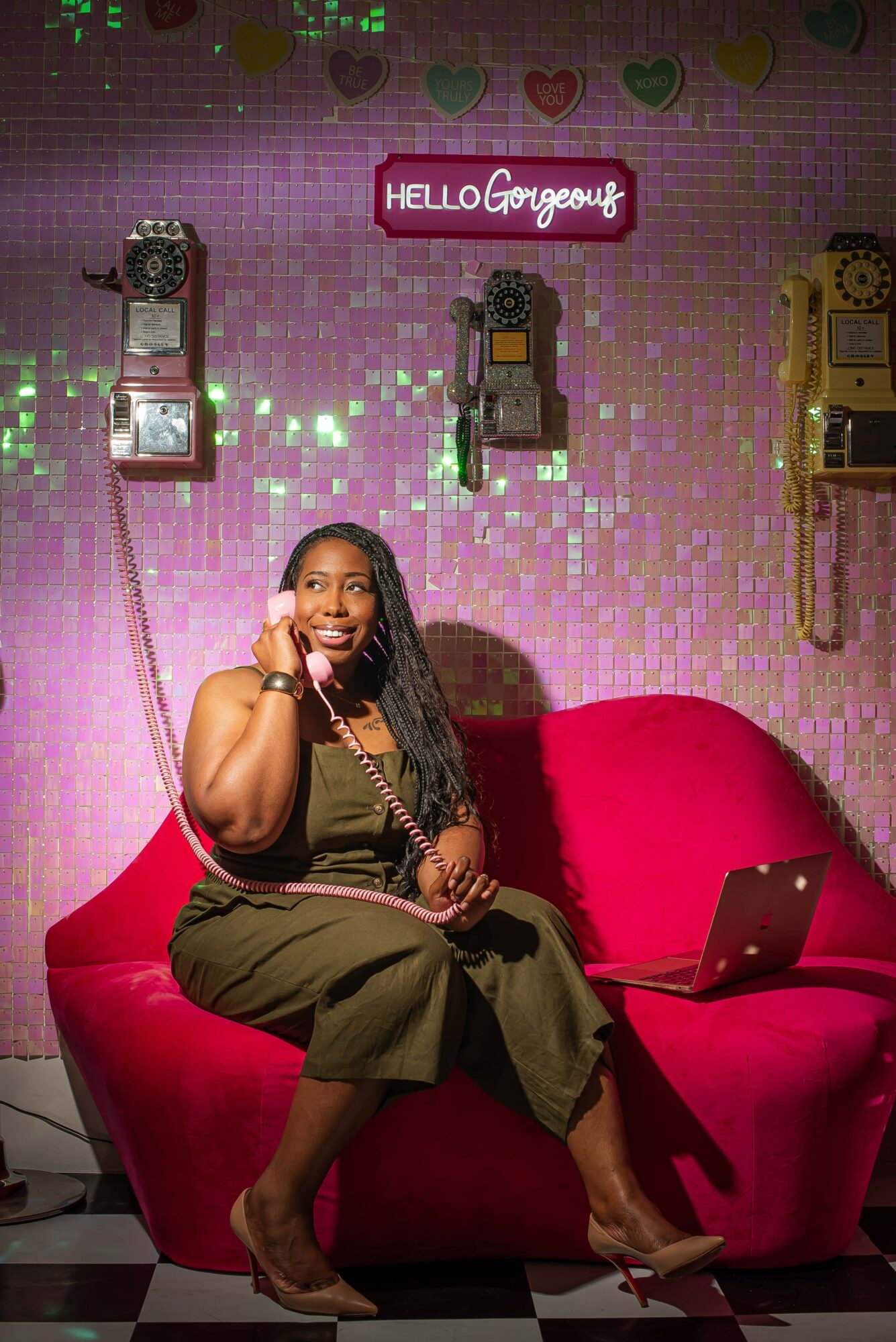 Woman sitting on a pink sofa with a laptop, smiling, against a pink wall with vintage phones and a sign that says 'HELLO Gorgeous'.