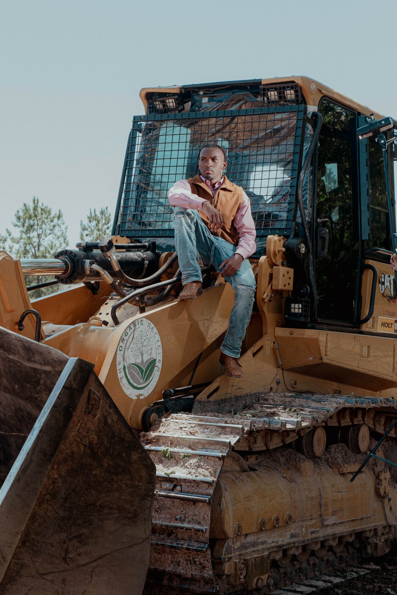 Person sitting on the edge of a large construction vehicle, wearing a brown jacket and jeans, with a clear sky background.