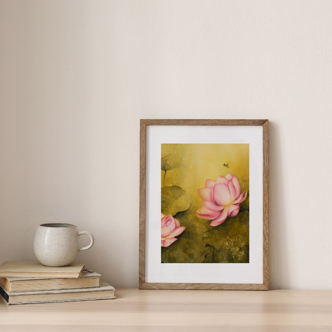 Framed watercolor painting of pink lotus flowers on a green background, placed on a light-colored surface with a mug and books nearby.