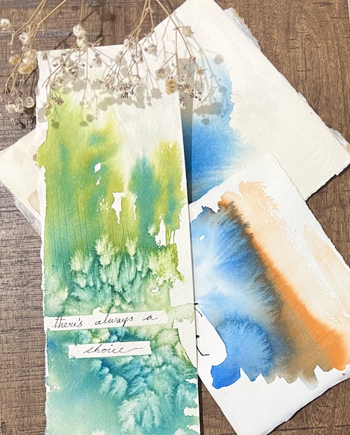 Two watercolor paintings with green, blue, and orange hues on torn paper, with dried flowers above, on a wooden surface.