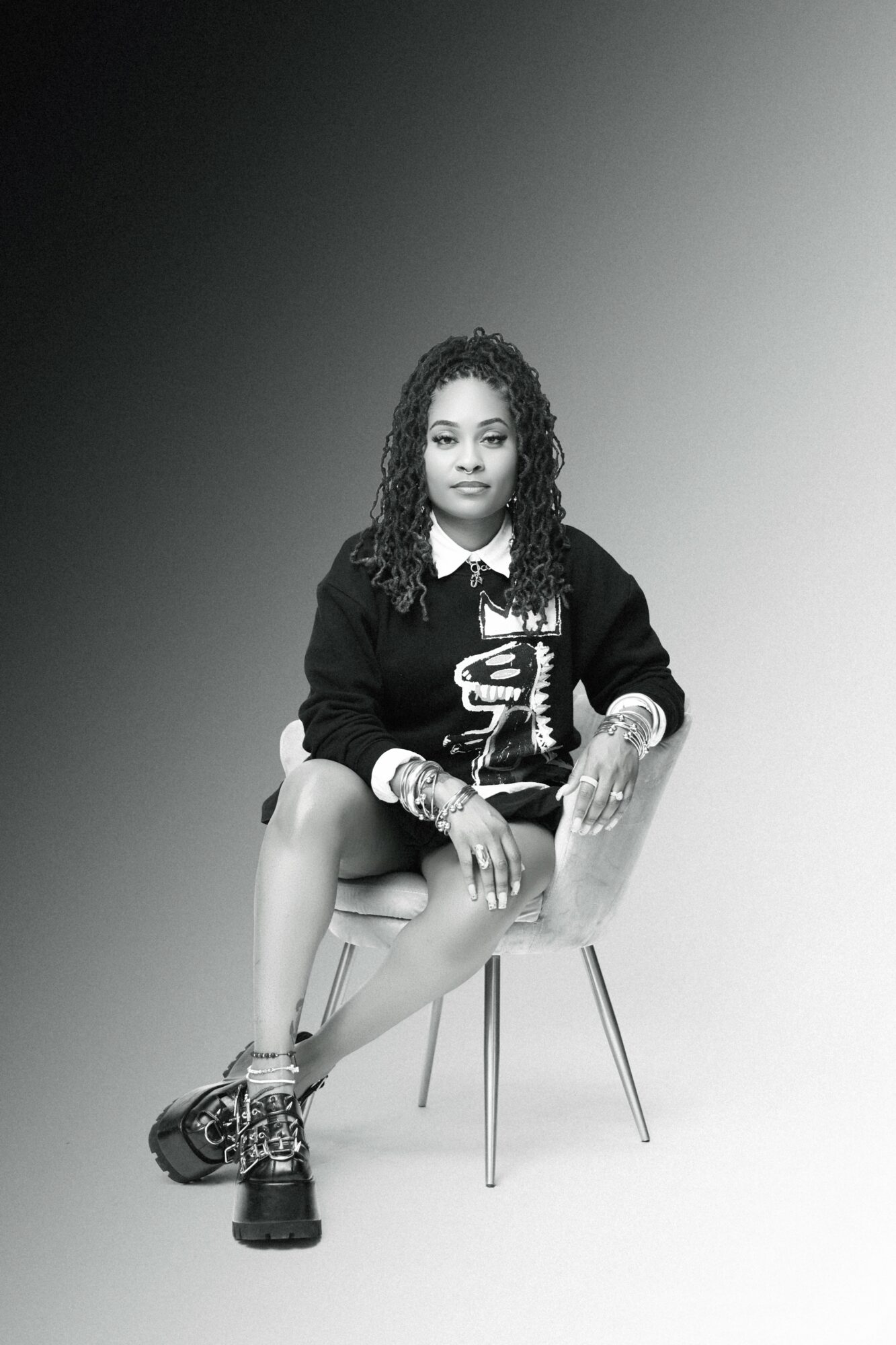 Young woman with curly hair sitting on a chair, wearing a dark sweater, shorts, and sneakers, holding a phone.