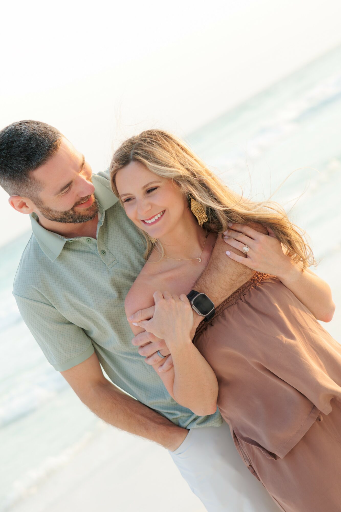 A smiling couple embracing on the beach, with the ocean in the background.