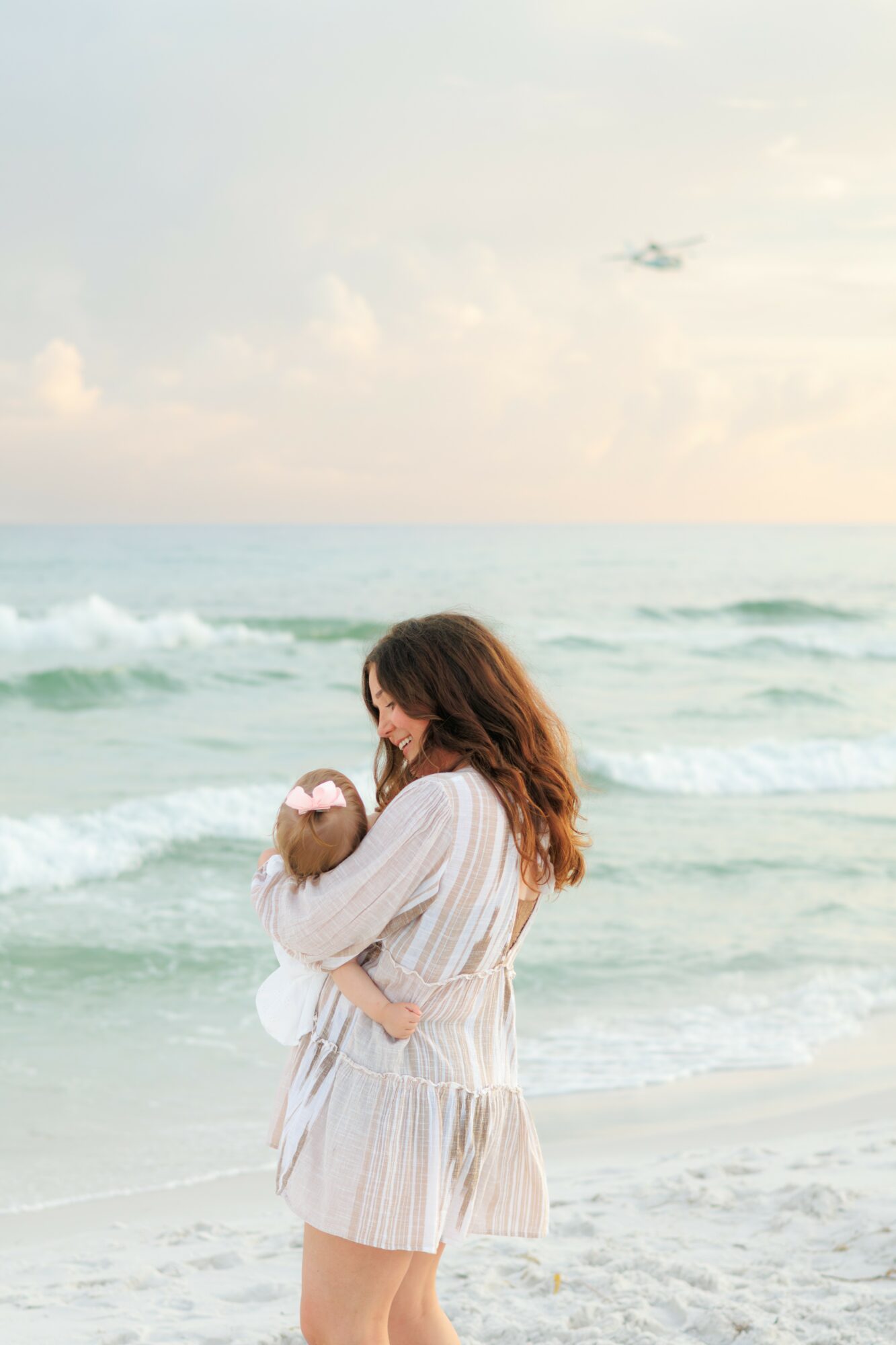 Woman holding a baby on a beach with ocean waves and sky in background.