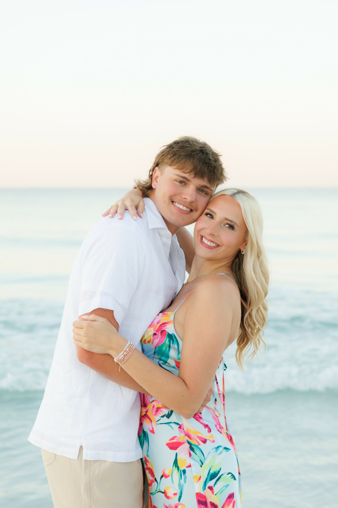 A smiling couple hugging on the beach with ocean waves in the background, woman in a colorful dress, man in white shirt.