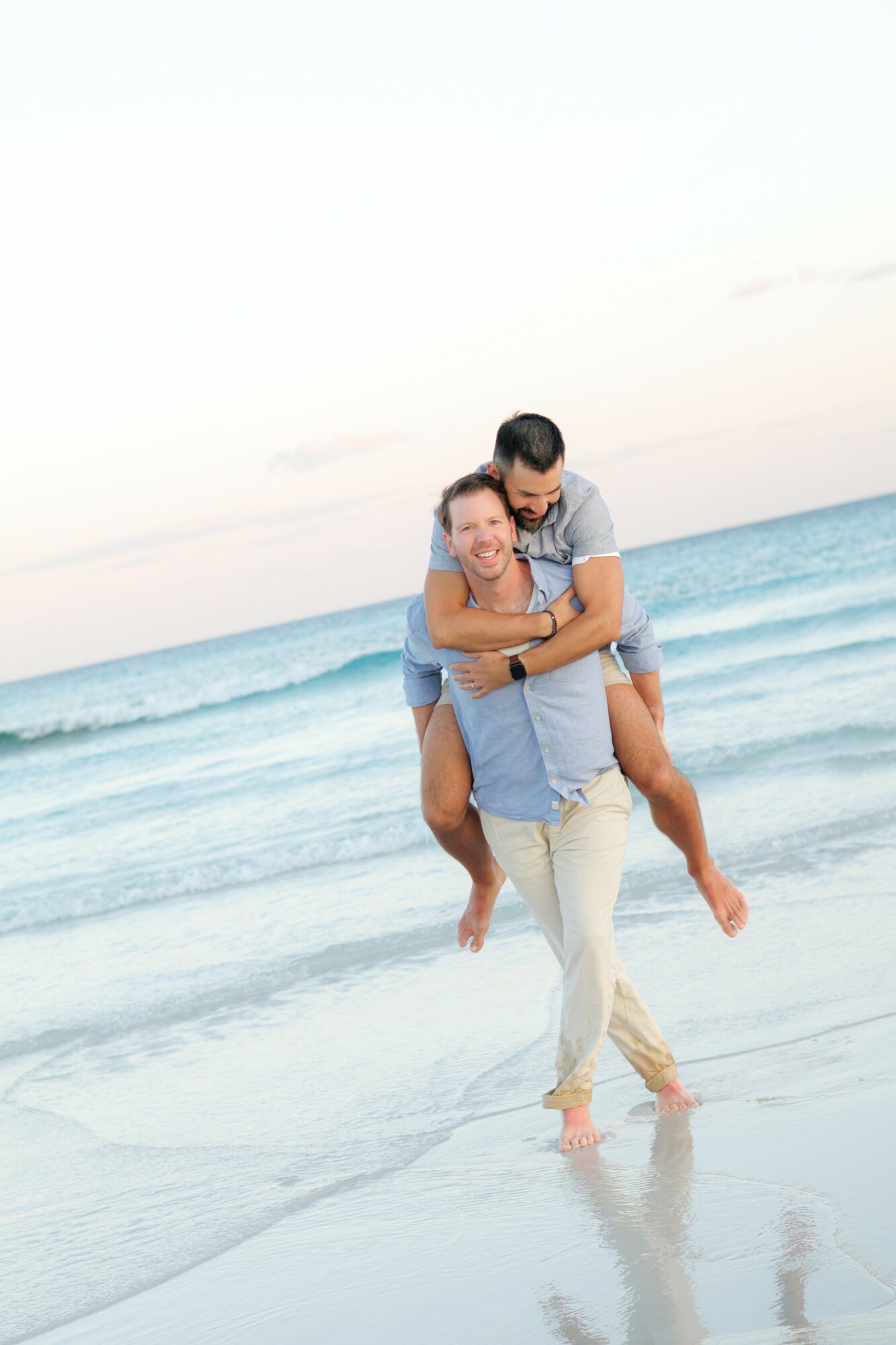 A couple on the beach, with the man giving the woman a piggyback ride, ocean waves in the background.