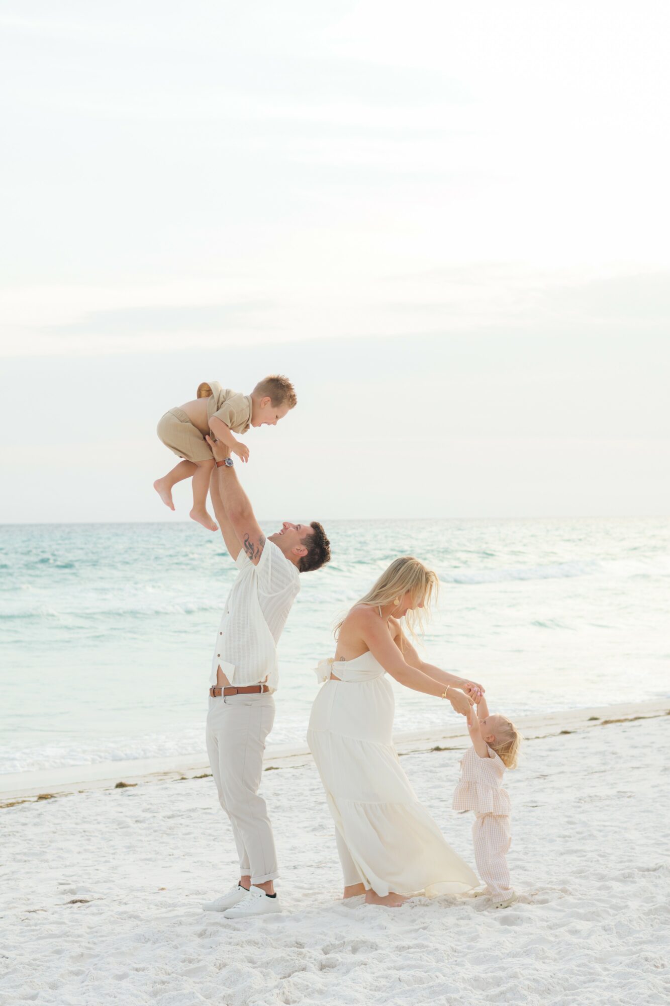 Family on the beach, a man lifts a child, a woman and a young girl play in the sand, ocean in background.