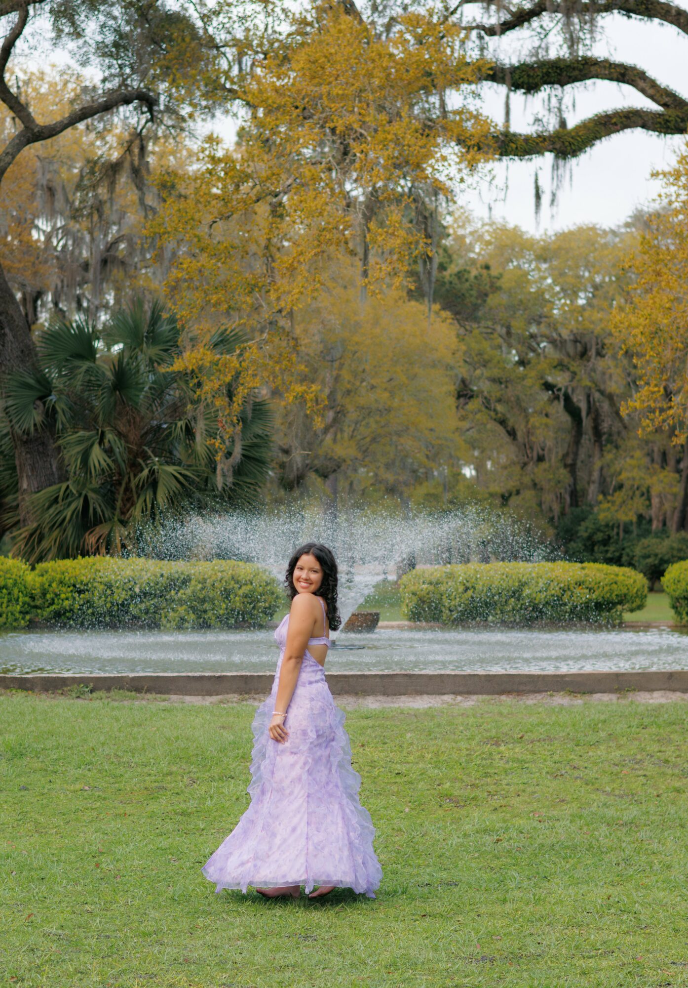 Woman in a lavender gown standing on grass in a park with trees and a fountain in the background.
