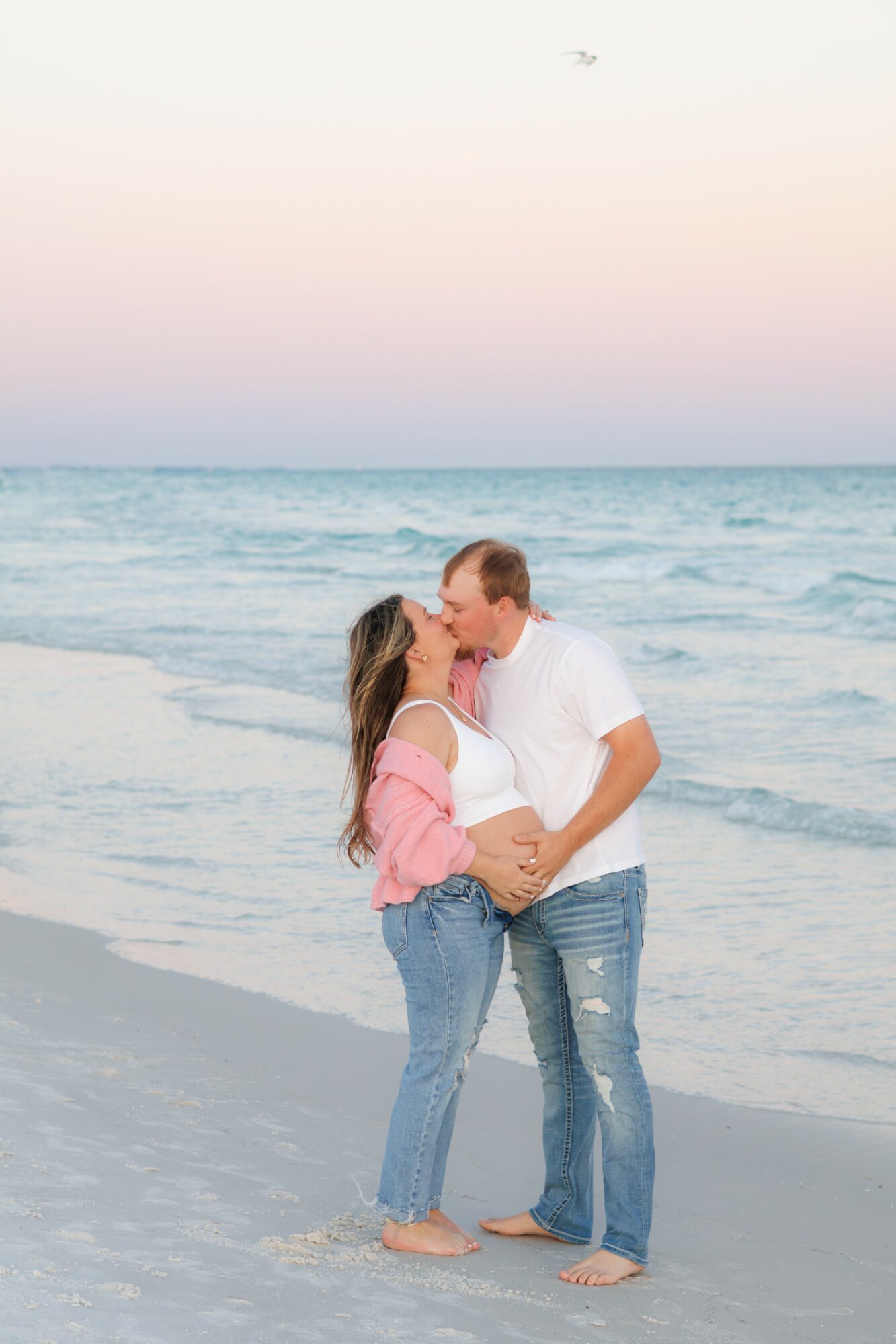 Couple kissing on the beach at sunset, holding a pregnant belly, ocean waves in background, pastel sky, barefoot, casual clothing.