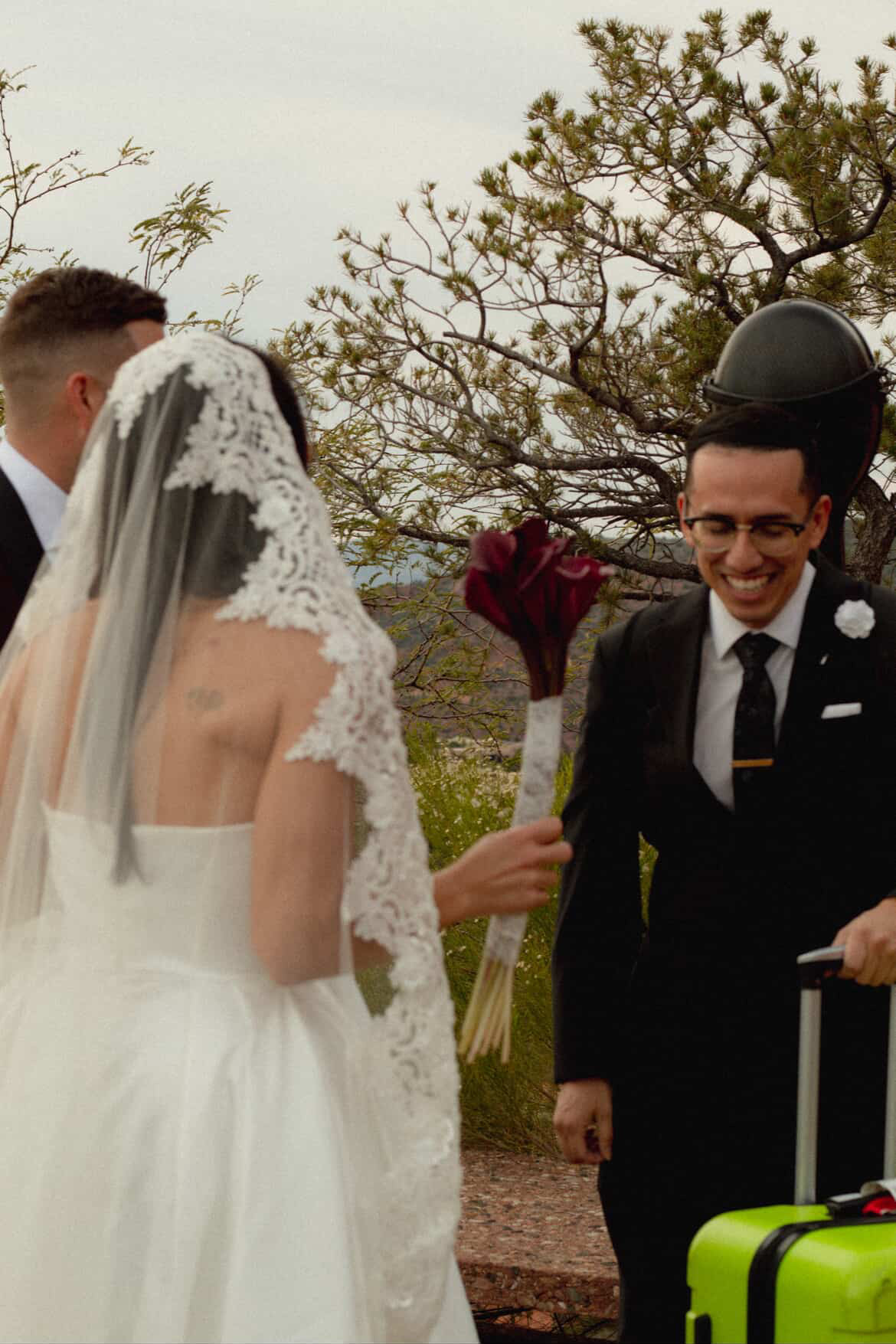 Bride and groom outdoors, bride holding bouquet, groom wearing helmet, trees in background, wedding attire, smiling.