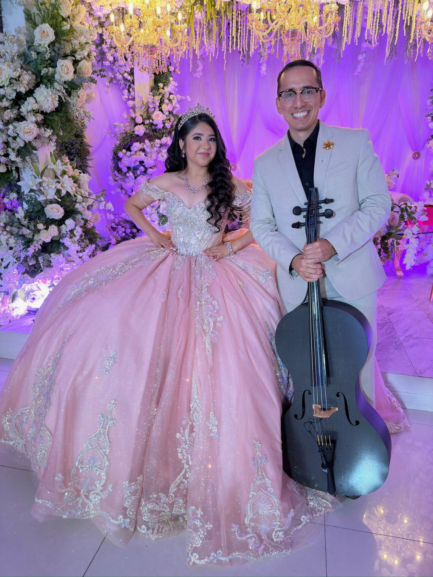 A woman in a pink ball gown and a man in a white suit with a cello pose together in front of a decorated backdrop.