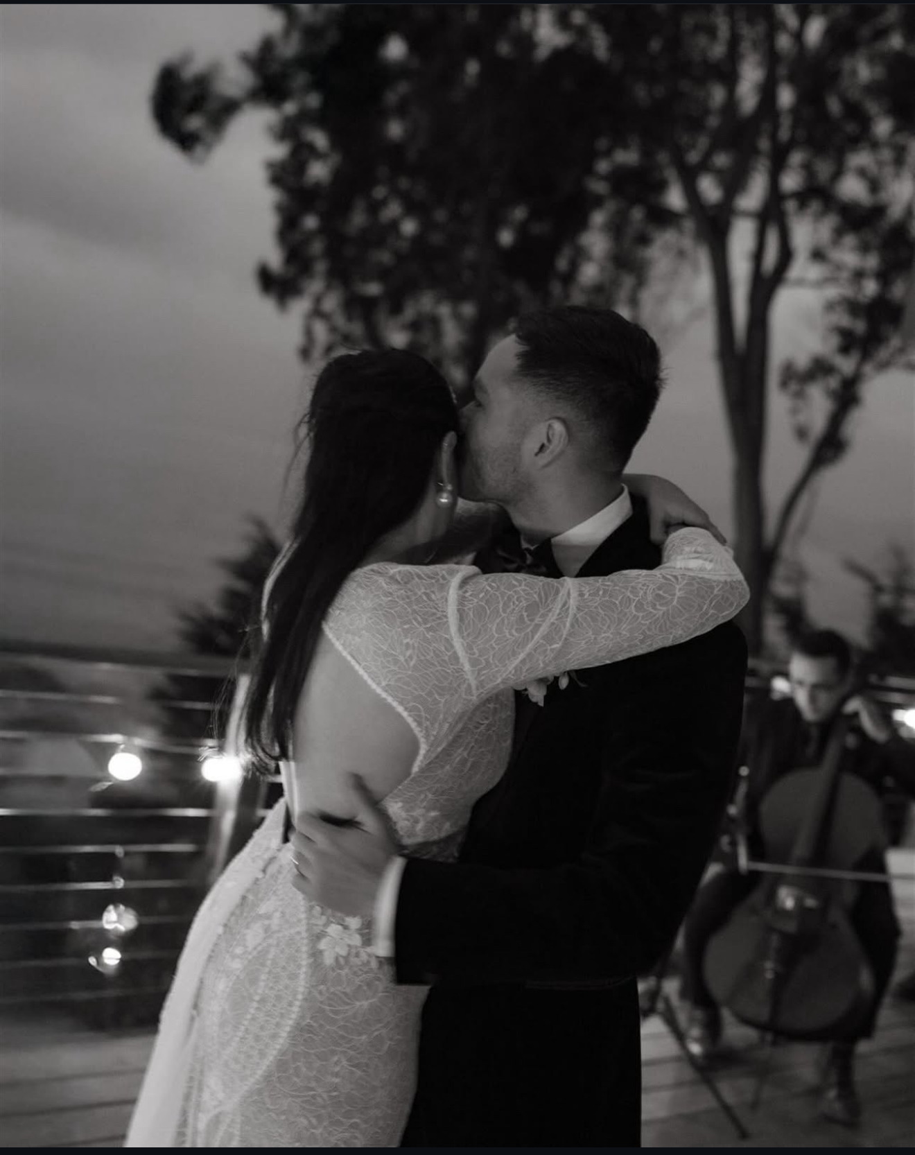 Couple kissing outdoors, woman in a dress and man in a suit, trees and people in background.