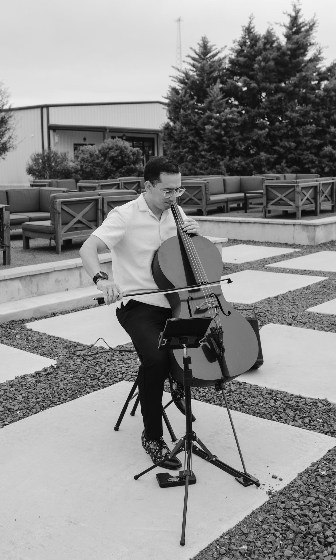 Person playing cello outdoors with trees and seating area in background, standing on a paved surface.